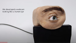 A realistic human eye with fake flesh and eyebrows shaped like a tube sits atop a monitor with a cable leading from it. The text reads We developed a webcam looking like a human eye