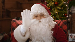 Santa is waving to the camera with a Christmas tree behind him.