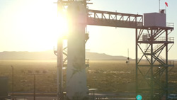 A rocket ship is on a launchpad with the bright Sun near the horizon behind it.