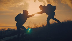 A closeup taken around dusk with thin white clouds shows a person in shadow wearing a backpack helping another person in shadow wearing a backpack up a grassy hill. The setting sun is directly behind where their hands meet