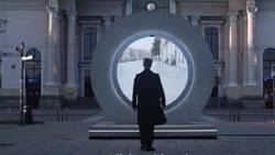 A view from behind and slightly to the left of a person in a long black coat standing in front of human-sized round display surrounded by stone that shows another location. It looks like the person could walk through to the other place.