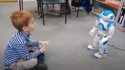 A child with red hair about 4 years old sits cross-legged on a carpeted floor of a classroom and is looking eye-to-eye at a small blue and white humanoid robot NAO about 3 feet away.