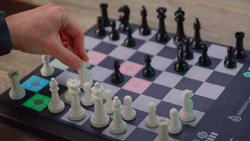 A closeup of a small chess board. A person is picking up a knight piece. The board is lighting up possible moves.