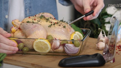 A closeup of a whole chicken in a glass dish with parsley, lemon slices, olives, quartered onion, and garlic cloves sitting on a cutting board. A person is inserting a probe that resembles an eyedropper about 4 inches long.