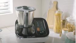 A black and silver device sits on a white kitchen counter. To the right is a cutting board, and clear jars of pasta, tall wooden salt and pepper shakers. The device has a short blender style container on the left and a touch screen display on the right.