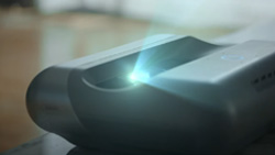 A closeup of a gray video projector sitting on a table. It has a bright light emminating from the center.