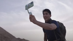 A person is hiking with a backpack on and taking a selfie with a smart phone attached to a compact phone stabilizer