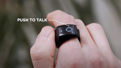 A closeup of the back of a person's hand with the thumb pressing on a button on a black ring with a display on the index finger. Text that reads Push to Talk has a line the points to the ring. The display shows a small circle made of dots.