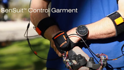 A closeup of a person outdoors with the left hand grasping the right wrist wearing black velcro straps on each forearm and over the back of the wrist that have wires attached. The person is holding two joystick controllers. The text reads SenSuit Control Garment.