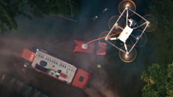 A view from above a hovering white drone with 6 propellers in a rectangle formation that is tethered to an emergency vehicle with a red cord