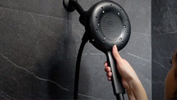 A closeup of a person in the shower with dark gray marble tile grabbing the handle of a black showerhead that is shaped like a magnifying glass