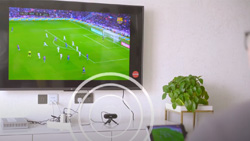 A television mounted on a wall with a desk underneath shows a small, black, pill-shaped device on a tripod. Concentric circles are overlayed to represent a transmitted signal